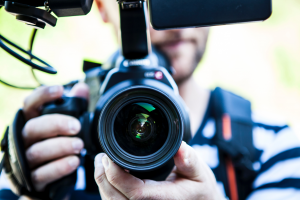 Ein Mann hält eine Fotokamera in der Hand, der Betrachter des Bildes sieht direkt in das Objektiv