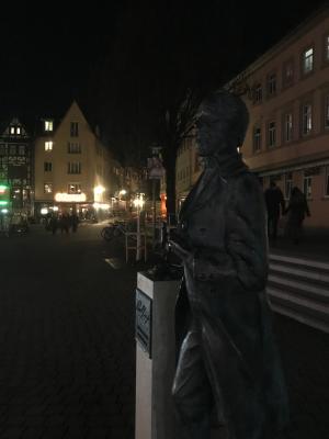 Ein Denkmal eines Mannes auf einem dunklem Platz, im Hintergrund Laternen einer Straße