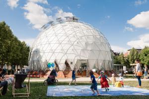 Kinder spielen in einem Park vor einem kuppelförmigen Pavillion.