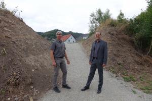 Bürgermeister Christian Gerlitz und KSJ-Mitarbeiter Jürgen Henning stehen auf einem geschotterten Weg. Der Weg führt durch einen Einschnitt in einem Lärmschutzwall. Im Hintergrund sind Häuser zu sehen.