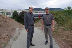 Bürgermeister Christian Gerlitz und KSJ-Mitarbeiter Jürgen Henning stehen auf einem geschotterten Weg. Der Weg führt durch einen Einschnitt in einem Lärmschutzwall. Im Hintergrund fährt ein Zug vorbei.