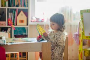 Kleines Mädchen sitzt an einem tisch in einem Raum mit Spielzeugen und schneidet etwas aus