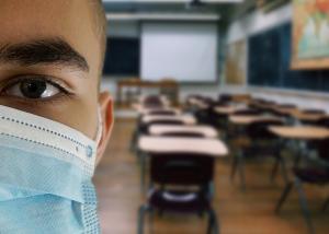 Halbes Gesicht einen Schülers mit Maske dahinter ein leeres Klassenzimmer