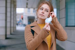 Eine junge Frau steht vor einem Gebäude und befestigt gerade eine Mund-Nasen-Bedeckung an einem ihrer Ohren