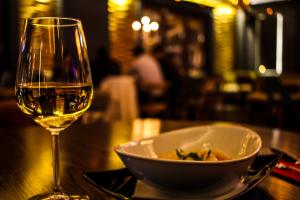 Ein Weinglas und ein Teller mit Essen auf einem Tisch in einem angenehm dunklen Raum, im Hintergrund sitzen Mneschen an einem Tisch, die unscharf zu sehen sind