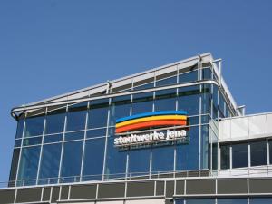 Blick auf eine Fassade mit vielen Glasflächen und Metall, im Hintergrund blauer Himmel und auf der Fassade drei farbige Bögen übereinander, darunter der Schriftzug Stadtwerke Jena