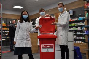Drei Menschen stehen vor einer Box, in welche sie Medikamentenverpackungen werfen. Im Hintergrund befinden sich viele Regale.