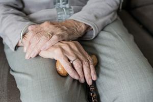 Blick auf Hände und Beine einer älteren sitzenden Person die einen Stock in der einen Hand hält