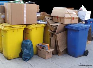Ein Haufen Kartons steht neben, auf und zwischen gelben und blauen Abfalltonnen.