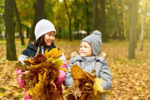 Es ist Herbst: Zwei Kinder lächeln sich an, sie tragen Mütze und beide eine Handvoll Laubblätter.