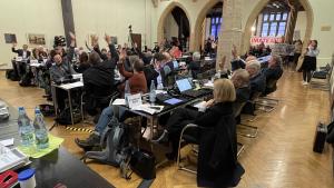 Abstimmung des Jenaer Stadtrats: Die meisten Stadträte heben die Hand. Im Hintergrund halten Personen Transparente hoch und fordern den Klima-Aktionsplan zu beschließen.