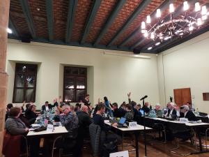 Stadtratsmitglieder sitzen an Tischen im Historischen Rathaus und heben die Hände, weil sie für einen Beschluss stimmen