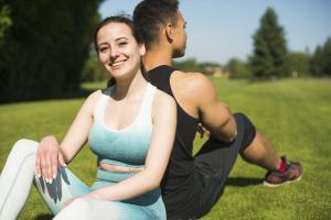 Eine junge Frau und ein junger Mann sitzen Rücken an Rück auf einer Wiese im Park. Sie tragenSportoutfits. Die Frau lächelt in die Kamera.
