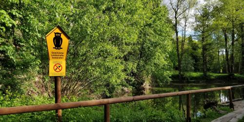 Vor einem Teich steht ein Schild. Auf dem Schild ist eine Eule abgebildet. "Geschützter Landschaftsbestandteil" ist zu lesen.