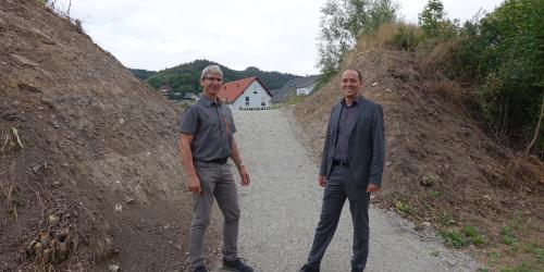 Bürgermeister Christian Gerlitz und KSJ-Mitarbeiter Jürgen Henning stehen auf einem geschotterten Weg. Der Weg führt durch einen Einschnitt in einem Lärmschutzwall. Im Hintergrund sind Häuser zu sehen.