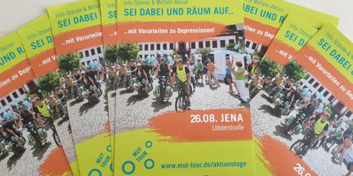 Mehrere Flyer der MUT-Tour liegen aufgefächert auf einem Tisch. Es ist ein Bild mit Fahrradfahrern zu sehen und der Termin in Jena.