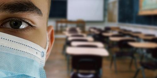 Halbes Gesicht einen Schülers mit Maske dahinter ein leeres Klassenzimmer