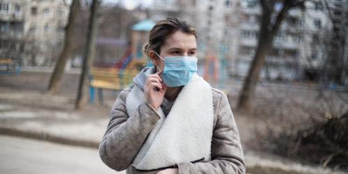 Frau mit Mund-NasenBedeckung steht vor Häuserblock