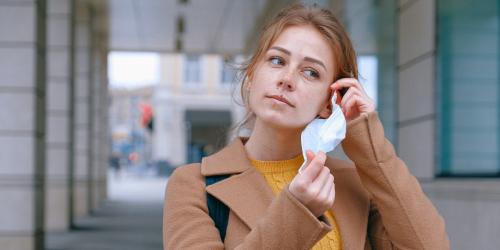 Eine junge Frau steht vor einem Gebäude und befestigt gerade eine Mund-Nasen-Bedeckung an einem ihrer Ohren