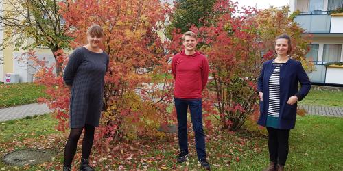 Zwei Frauen und ein Mann stehen im Herbst vor einem Busch.
