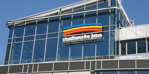Blick auf eine Fassade mit vielen Glasflächen und Metall, im Hintergrund blauer Himmel und auf der Fassade drei farbige Bögen übereinander, darunter der Schriftzug Stadtwerke Jena