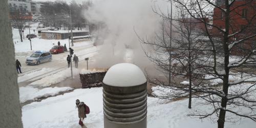 Blick auf eine verschneite Straße mit einer Rauch- bzw. Wasserdampfwolke, mehreren Fußgängern und Fahrzeugen, unter anderem eine