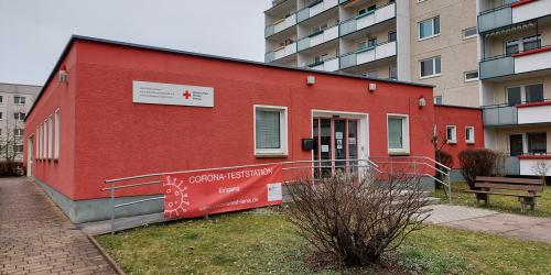 Ein rotes gebäude steht im Vordergrund, an einem Geländer befindet sich ein Plakat mit der Aufschrift Corona-Teststation, im Hintergrund ein großer Wohnblock in grau mit vielen Etagen.