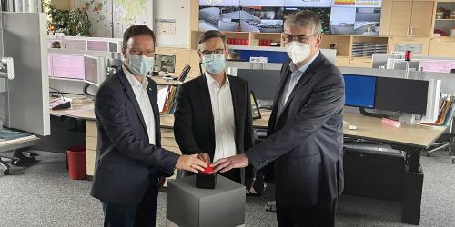 Drei Männer in einem Raum voller Computerarbeitsplätze drücken auf einen großen Knopf
