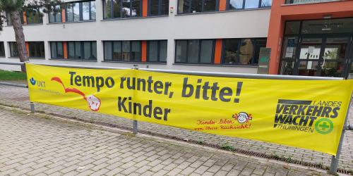Blich auf eine Schule. An einem Geländer hängt ein großes gelbes Plakat mit der Aufschrift: Tempo runter, bitte! Kinder