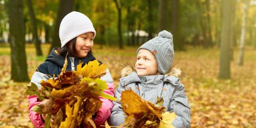Es ist Herbst: Zwei Kinder lächeln sich an, sie tragen Mütze und beide eine Handvoll Laubblätter.