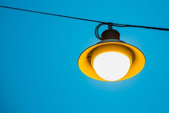 Straßenlampe hängt an einem Seil und leuchtet, dahinter blauer Himmel.