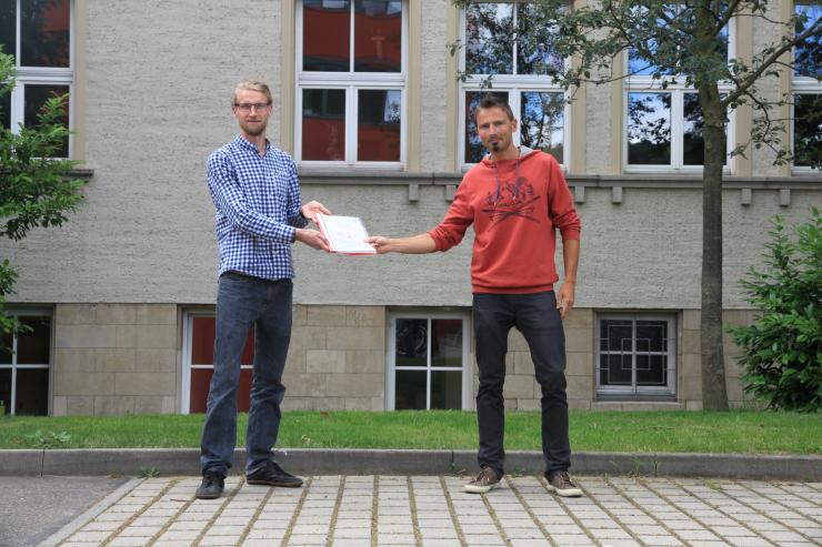 Zwei Personen stehen vor einem Gebäude. Die eine Person übereicht ein Dokument der anderen.