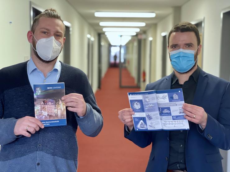 Herr Heise und Her Koppe stehen im Gang eines Verwaltungsgebäudes und präsentieren beide die Jubiläumsbroschüre.