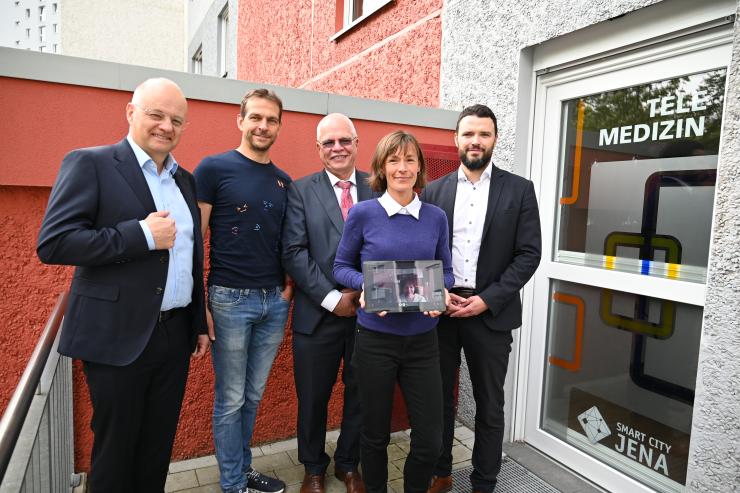 Vier Männer und eine Frau stehen vor dem Eingang eines Telemedizinraums. Die Frau hält ein Tablet, auf der eine weitere Frau zu sehen ist.