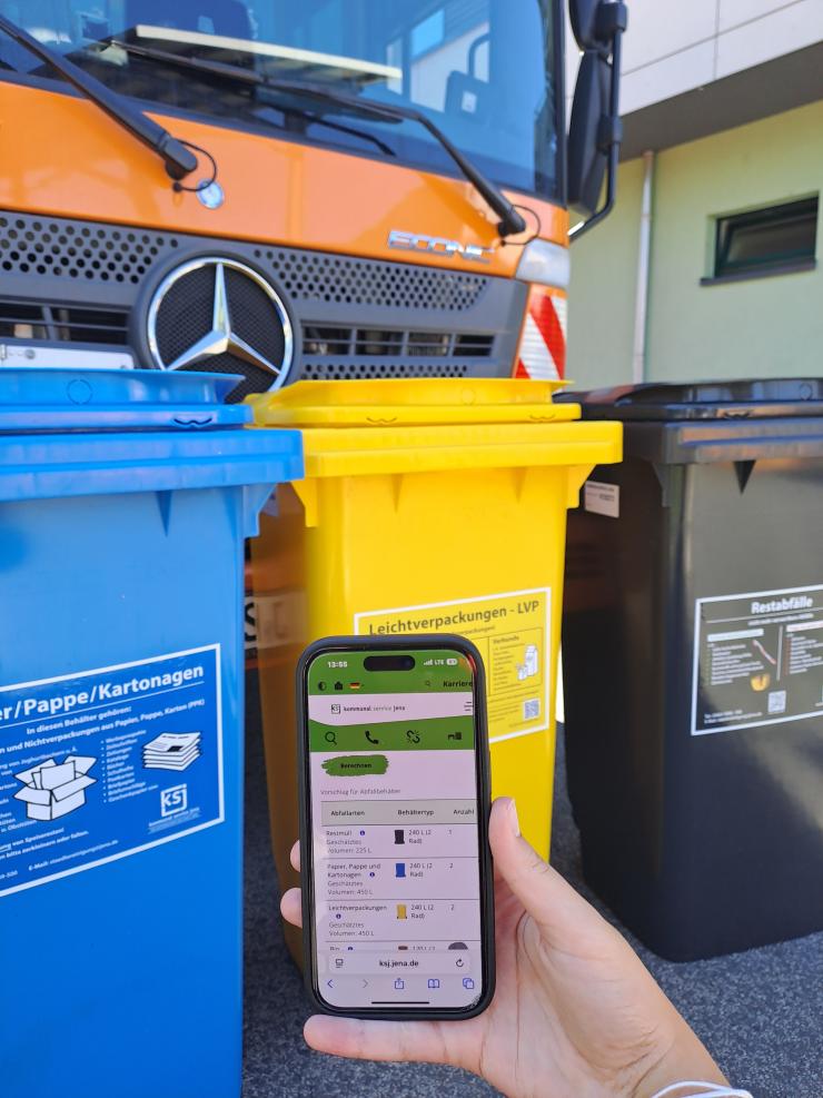 Handy mit abgebildeten Behälter-Konfigurator vor einer blaueb, gelben und scharzen Mülltonne sowie einem Müllfahrzeug