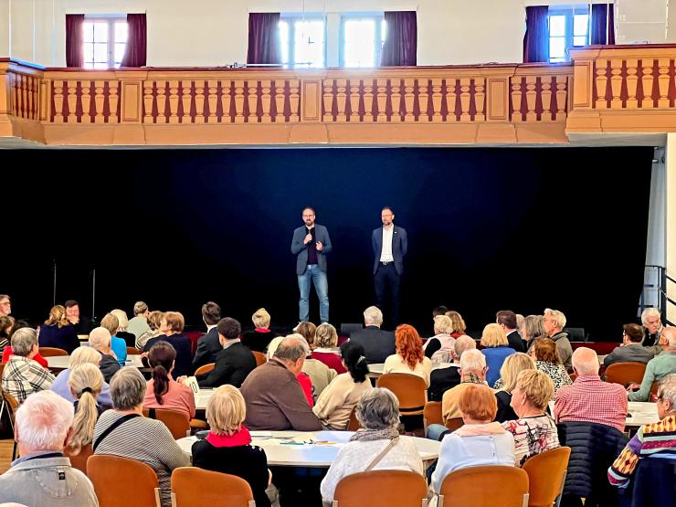 Zwei Männer stehen auf einer Bühne. Davor sitzen Menschen an runden Tischen