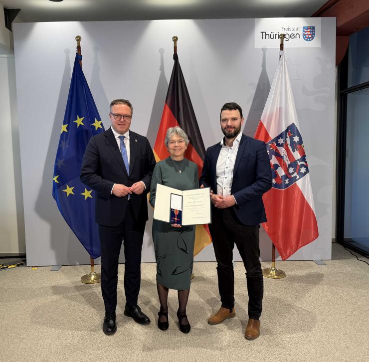 Drei Menschen stehen vor Fahnen und schauen in die Kamera.