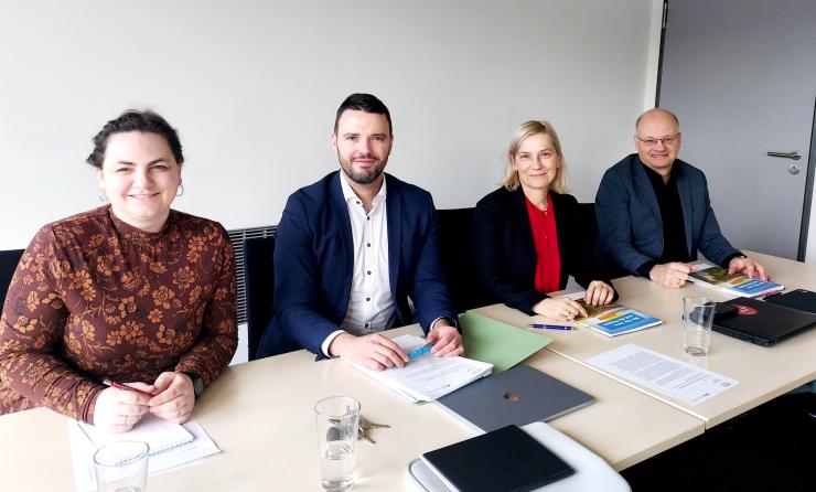 Zwei Frauen und zwei Männer sitzen an einem Tisch, vor ihnen sind Unterlagen ausgebreitet, sie lächeln in die Kamera.