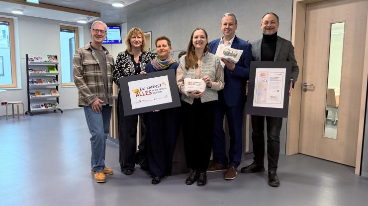 Sechs Personen stehen in einem modernen Innenraum zusammen und präsentieren gerahmte Plakate sowie kleine Taschen; einige halten Produkte in den Händen.