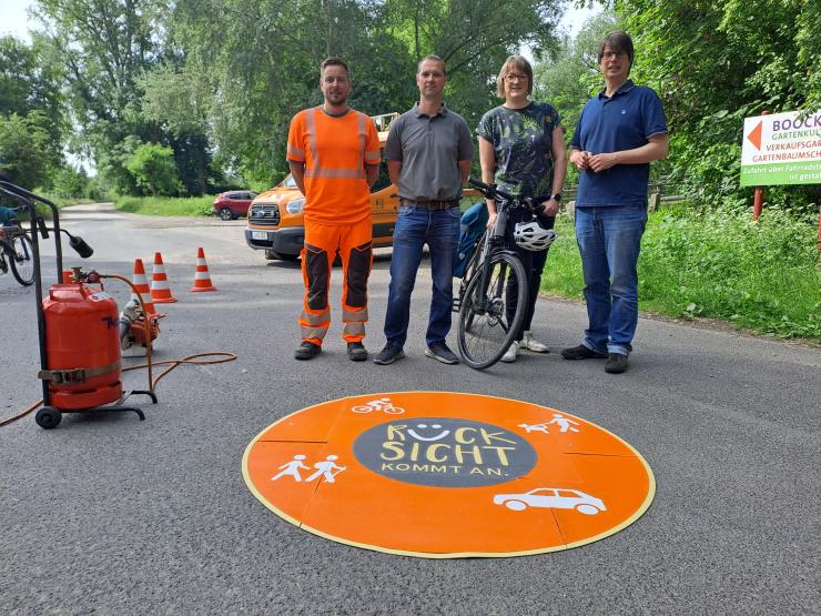 Drei Männer und eine Frau mit Fahrrad stehen hinter einer runden Markierung auf der Straße.
