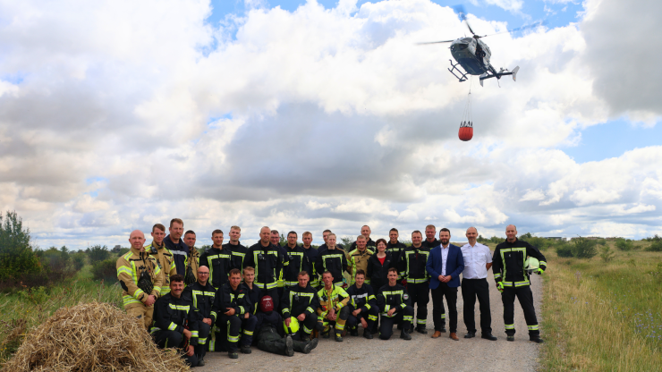 Mehrere Männer stehen auf einem weiten Wiesenfeld, ein Hubschrauber im Hintergrund.