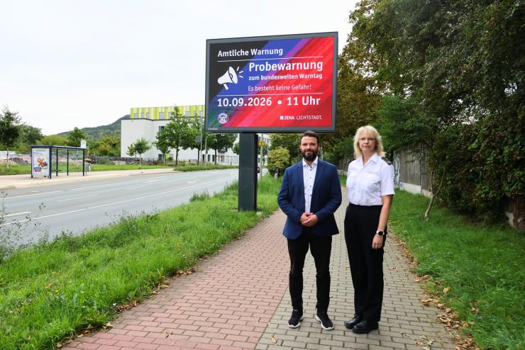 Ein Mann und eine Frau stehen vor einer digitalen Anzeigetafel in Jena.