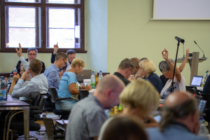 Eine Gruppe von Menschen bei einer Abstimmung des Stadtrates Jena im Rathaus. Die Personengruppe sitzt an Tischen, manche heben eine Hand zur Abstimmung.
