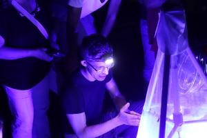 Ein Mann mit einer Stirnlampe hockt nachts vor einer Lichtanlage und erklärt etwas. Personen stehen um in rum. 