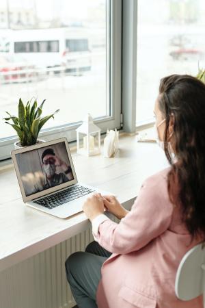 Videochat, eine Person ist mit Mundschutz auf dem Blidschirm eines Laptop zu sehen die andere Person sitzt davor