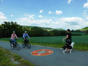 Drei Menschen auf einem Fahrradweg, zwei mit Fahrrad, eine Dame mit Hund