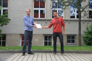 Zwei Personen stehen vor einem Gebäude. Die eine Person übereicht ein Dokument der anderen.