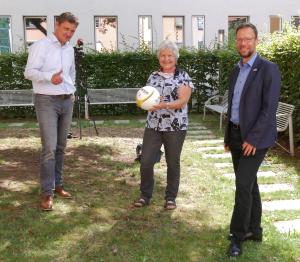 Drei Mensch schauen in die Kamera, die Frau in der Mitte hält einen Fußball.