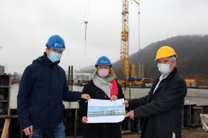 Drei Personen halten einen Bauplan in die Kamera. Sie stehen auf einer Baustelle.