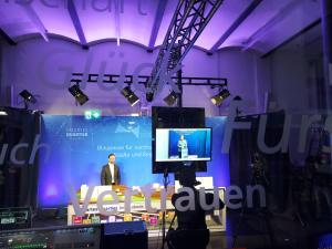 Ein Mann steht in einem Filmaufnahmestudio vor einem blauen Hintergrund. Rechts neben ihm sieht ist auf einem eine Frau zu sehen. Das Bild ist aus Perspektive des Technikpults aufgenommen worden.